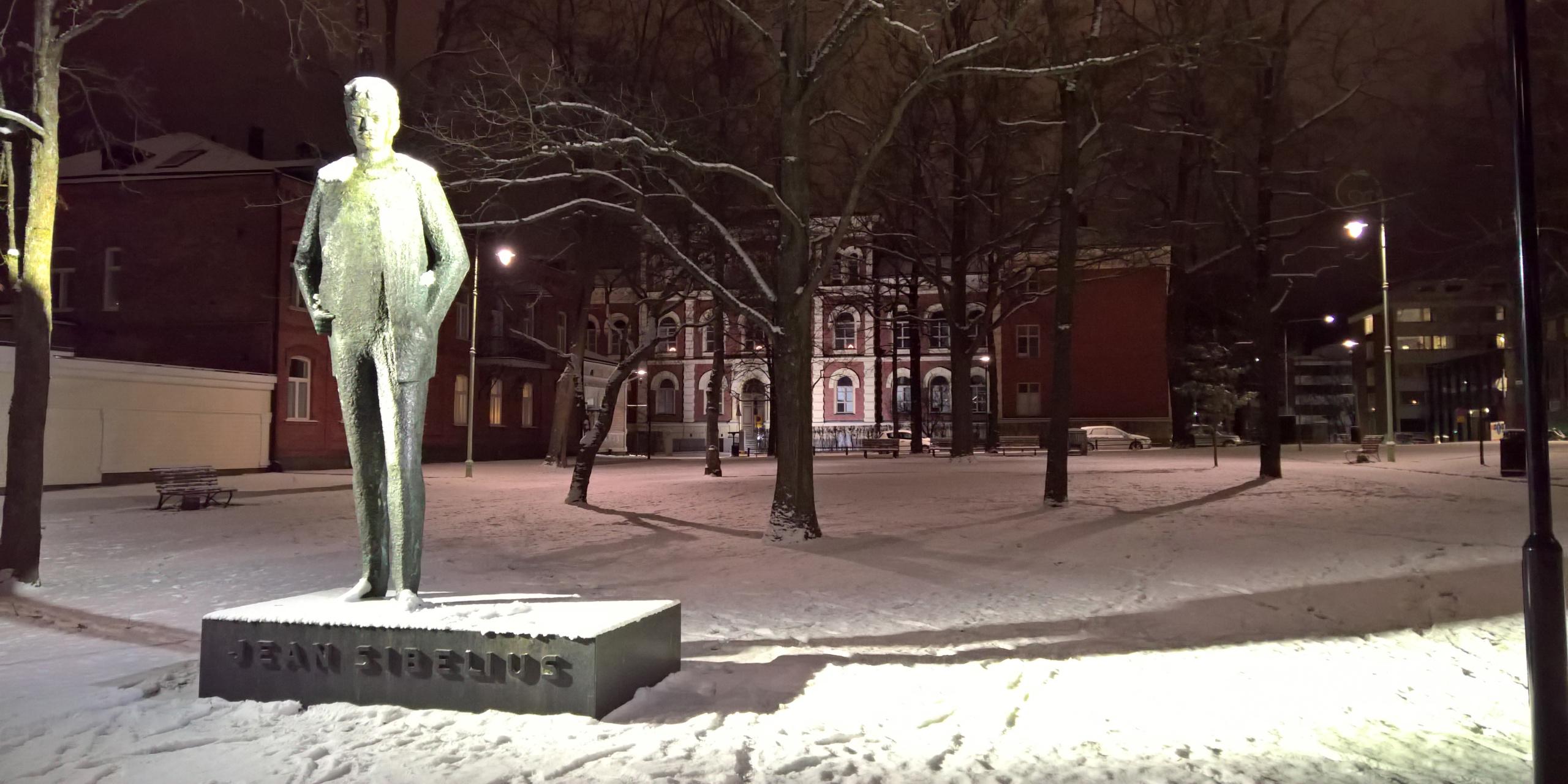 Nuorta Sibeliusta kuvaava pronssiveistos jalustallaan Hämeenlinnan Sibeliuksenpuistossa, talvisena iltana. Taustalla Hämeenlinnan Lyseon rakennus.
