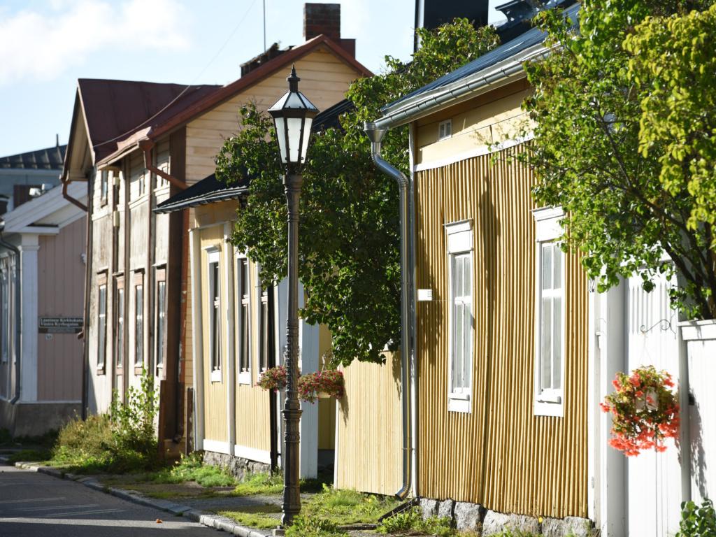 Kadun varressa lähikuvassa vanhoja puutaloja, Ruskean ja okran värisiä, osassa valkoiset ikkunapuitteet. Julkisivuista ja niitä jatkavista aidoista muodostuu umpikortteli, aitojen päälle talojen välistä pilkottaa puita. Kapea ruohottunut jalkakäytävällä, jossa vanha lyhtymallinen ja koristeellinen valaisintolppa. Kesäkukkiaroikkuu amppeleissa.