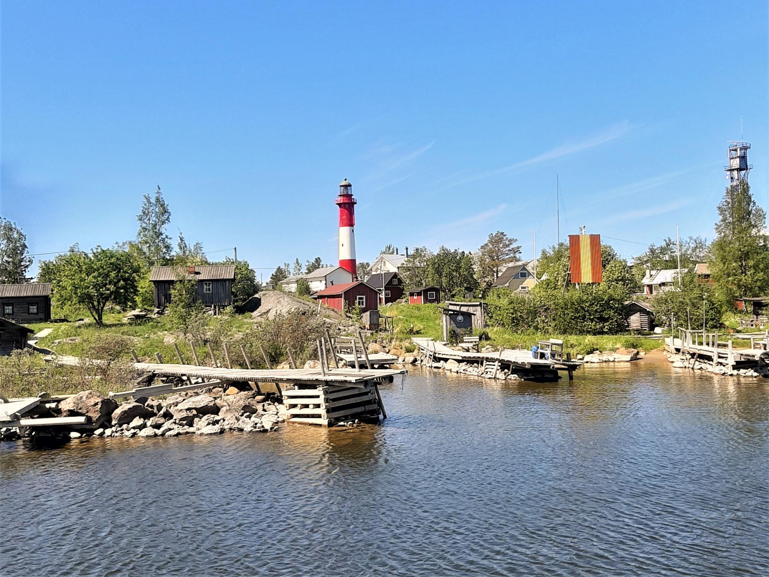 Vanha kalastajakylä kesällä, rantaviivassa kiviröykkiöille perustettuja vähän vinksottavia puulaitureita, taustalla rakennuksia ja keskellä kuvaa puna-valkoinen majakka.